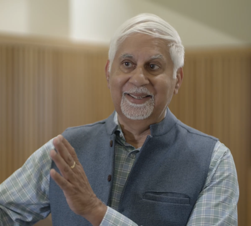 Vinit Nijhawan wearing a blue waistcoat and checked shirt, gesturing with both hands in an indoor setting.