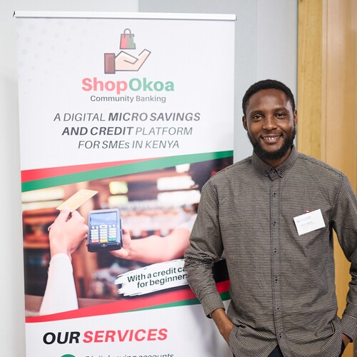 Kelvin stands next to a banner with the 'ShopOkoa' logo.